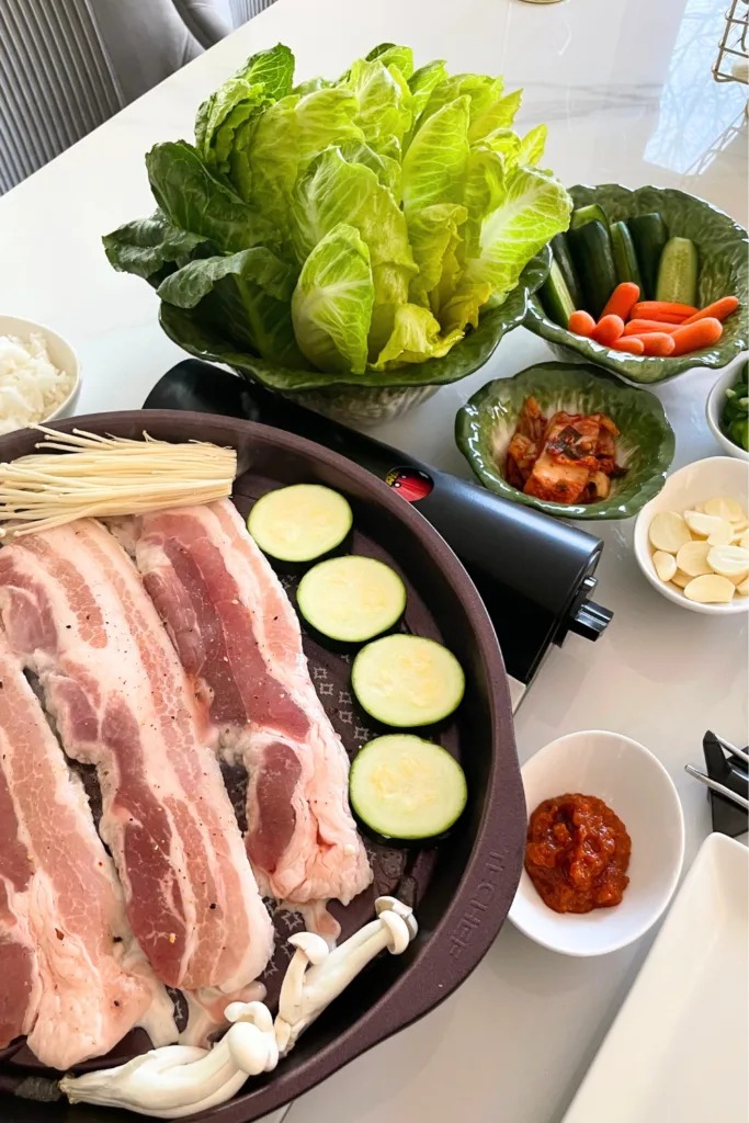 alt="Korean BBQ table setting with pork belly (pork belly) served with side dishes and lettuce wraps"