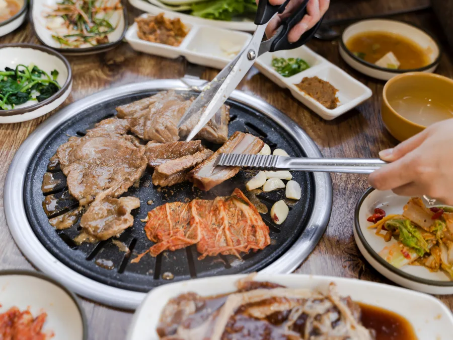alt="Using tongs and scissors to cut grilled meat into bite-sized pieces on a Korean BBQ grill"