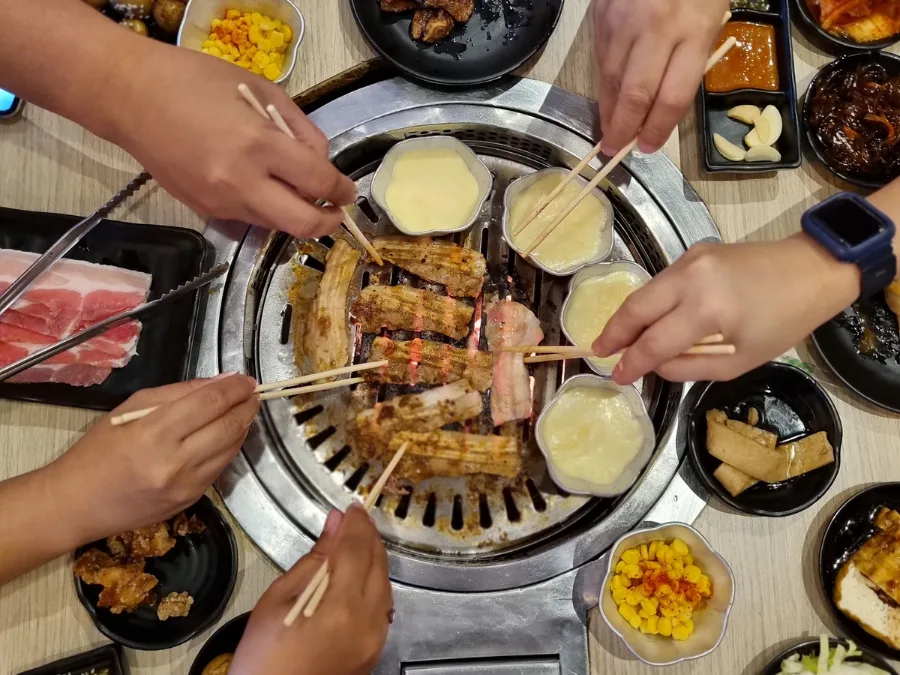 alt="Five people using chopsticks to pick up meat from a Korean BBQ grill"