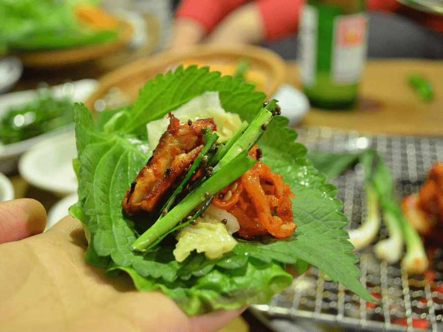 alt="Making a Korean BBQ wrap with lettuce, perilla leaves, kimchi, grilled meat, and chive salad"  
