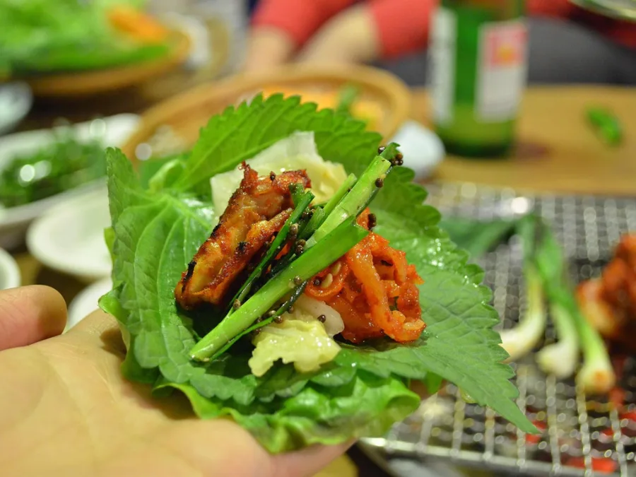 alt="Making a Korean BBQ wrap with lettuce, perilla leaves, kimchi, grilled meat, and chive salad"

