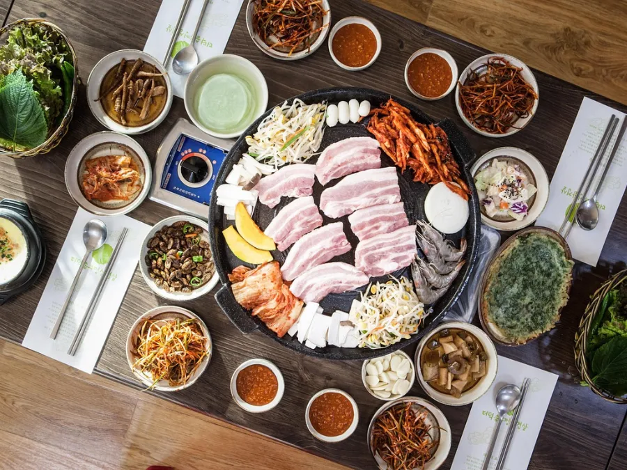 alt="Korean BBQ table setting with pork belly and a wide variety of side dishes"
